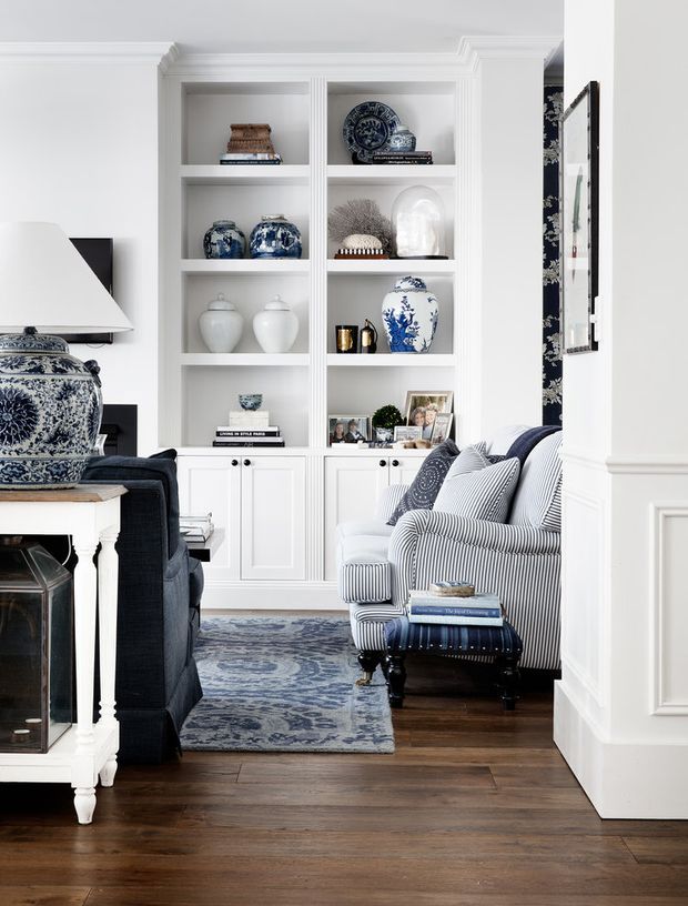 A refined traditional living room with white built-in display cabinetry, decorative moulding, dark timber flooring, and blue-and-white patterned furnishings and ceramics. A striped armchair and ottoman sit beside a dark sofa, while framed photographs and accessories fill the shelving. The room has a layered, classic look with strong navy, white, and timber accents.