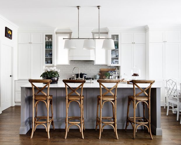 Bright, refined white kitchen with a large central island, four woven rattan bar stools, white shaker-style cabinetry, glass-front display cupboards, pale tiled backsplash, and a row of white pendant lights. Dark timber flooring and a grey island base add contrast, while a small dining area is visible at the right.