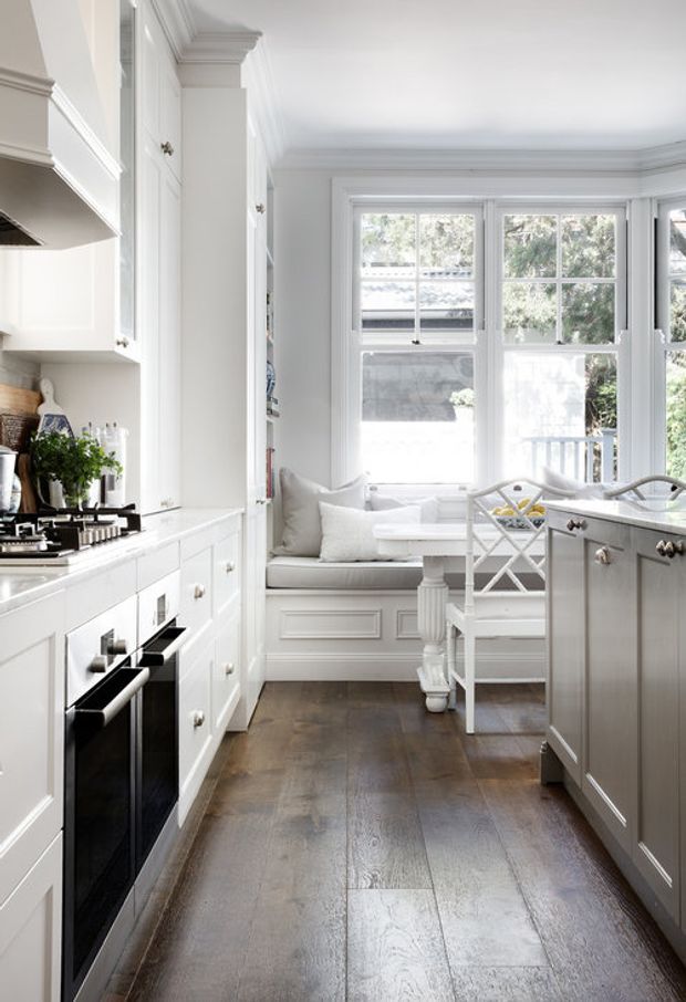 A bright, narrow traditional-style kitchen with white shaker cabinetry, a dark timber floor, integrated black appliances, and a central island with a pale grey finish. The room includes a built-in window seat with cushions, a small dining area, and large multi-pane windows bringing in abundant natural light. Decorative cornices, panelled cabinetry, and classic hardware create a refined, timeless character.