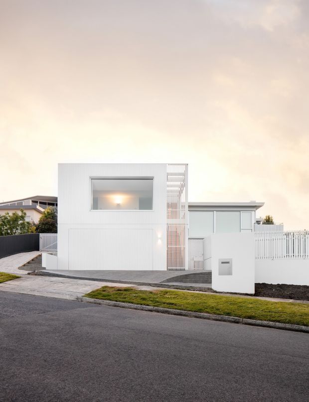 Street-facing exterior photograph of a contemporary white residential house. The facade combines vertically ribbed white cladding, a large square upper-level window, a narrow screened section, and a low flat-roofed wing with pale glazing. A white picket-style boundary fence, paved driveway, front utility box, and simple lawn verge are visible. The image has a soft, warm, overcast atmosphere.