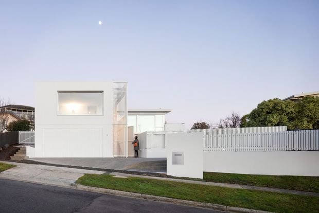Street-facing view of a minimalist white residential building with a boxy two-storey form, large horizontal window, vertical slatted screening, flat roof, and a matching white slatted boundary fence. A paved driveway and front entry are visible, with neighbouring houses and landscaping in the background.