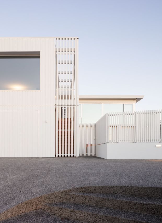 Minimalist contemporary residential facade in white, featuring a tall vertically screened entry or stair element, flush garage or service door, large dark-framed window, low horizontal roofline, and a white slatted boundary fence. The foreground includes a textured dark paved driveway or courtyard with curved bands, while the composition is clean and geometric under a clear sky.
