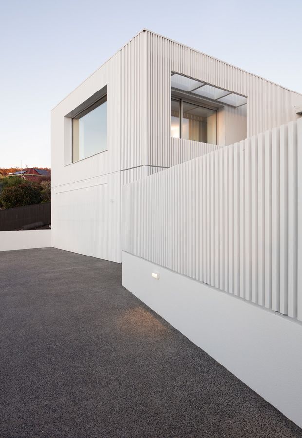 Exterior view of a minimalist contemporary residence with crisp white vertically ribbed cladding, large recessed glazed openings, and a matching privacy screen. The image is taken from a dark paved driveway or forecourt, with subtle wall lighting and neighbouring rooftops visible in the background.