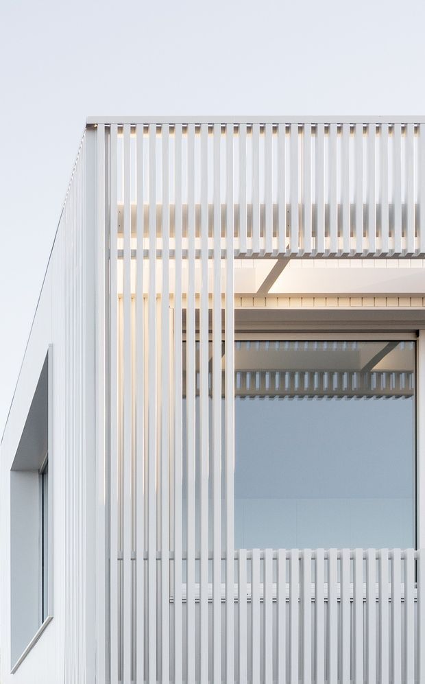 Close-up exterior view of a crisp white contemporary building facade with extensive vertical screening or balustrade fins. Large glazing, projecting structural elements, and layered open balconies create a highly geometric, light-filled composition.