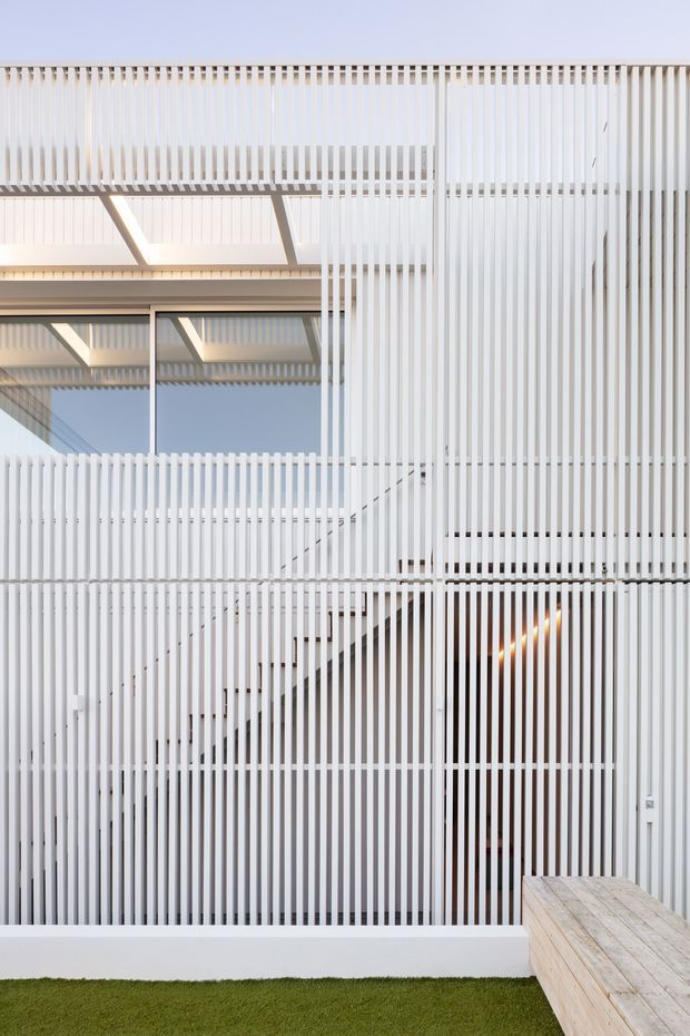 A bright contemporary multi-level building facade viewed from an outdoor deck or terrace. The facade is screened by closely spaced white vertical slats, creating a layered geometric rhythm and filtered views of glazing, stairs, and warm interior lighting. A timber deck edge and a strip of lawn occupy the foreground, while exposed roof or balcony framing is visible above.