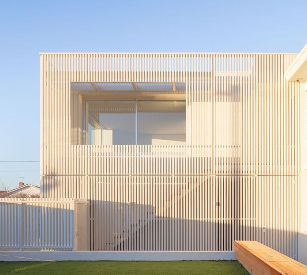 Contemporary two-storey residential facade viewed from outside, featuring a warm pale facade screened by closely spaced vertical timber battens. The battens create layered privacy and sun-shading around large upper-level glazing and a recessed balcony or terrace. A matching slatted gate and fence sit at street level, with a simple lawn and timber bench in the foreground under clear blue sky.