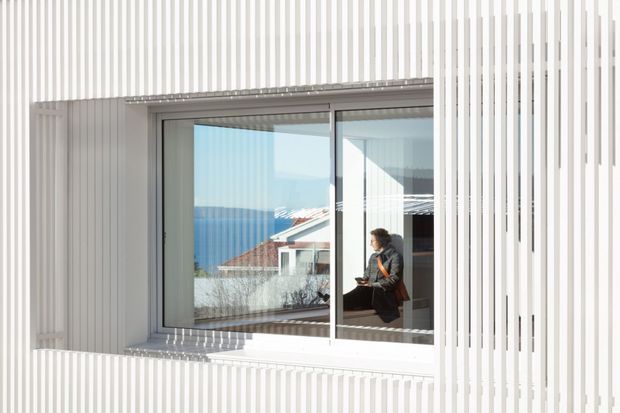 Exterior architectural view of a contemporary building facade clad in closely spaced vertical white slats. A large recessed sliding window provides a view into an interior with a seated person, while also reflecting or framing a neighbouring red-roofed building and blue water or sky beyond. The image emphasizes the rhythmic screen facade, deep window reveal, and strong natural light.