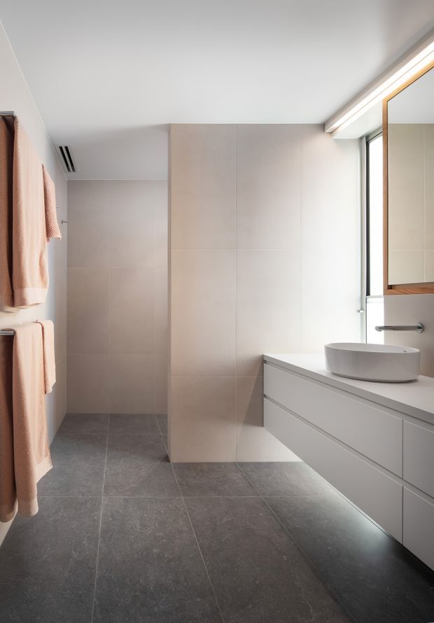 A bright, contemporary bathroom with a walk-in shower at the rear, large dark grey floor tiles, pale beige wall tiles, and a long white floating vanity with integrated drawers. A white vessel basin sits beneath a mirrored cabinet, with a narrow vertical window providing natural light. Warm beige towels hang on the left.