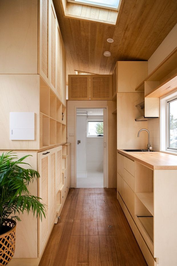 A narrow, compact galley kitchen with extensive light timber cabinetry, open shelving, woven-panel cabinet fronts, and a timber benchtop. A stainless-steel or chrome gooseneck tap sits beneath a large window, while a skylight brings natural light through the timber-lined ceiling. Timber flooring continues toward a doorway leading to a small tiled utility or bathroom area. A leafy plant and integrated storage add warmth and texture.