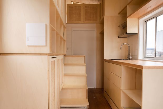 Compact, warm-toned galley kitchen with extensive light timber cabinetry and open shelving. A timber staircase rises beside the cabinetry toward a closed interior door, while the kitchen run includes a wood benchtop, inset sink, pull-out tap, drawers and open storage beneath a large window. The space is brightly lit by natural light and has dark timber flooring, creating a highly efficient, custom-built small-space layout.