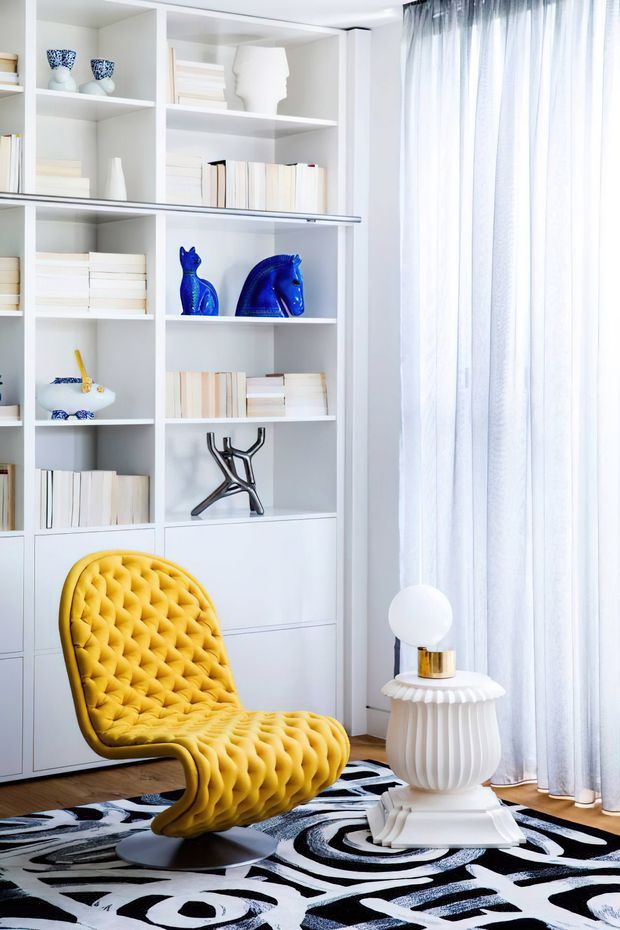 A bright, highly stylised living room corner featuring a built-in white shelving and cabinetry wall filled with books and sculptural objects. A vivid mustard-yellow tufted sculptural lounge chair sits on a bold black, white and grey abstract rug, beside a white fluted pedestal side table with a spherical lamp. Floor-to-ceiling sheer curtains provide soft daylight, with warm timber flooring visible beneath the furnishings.