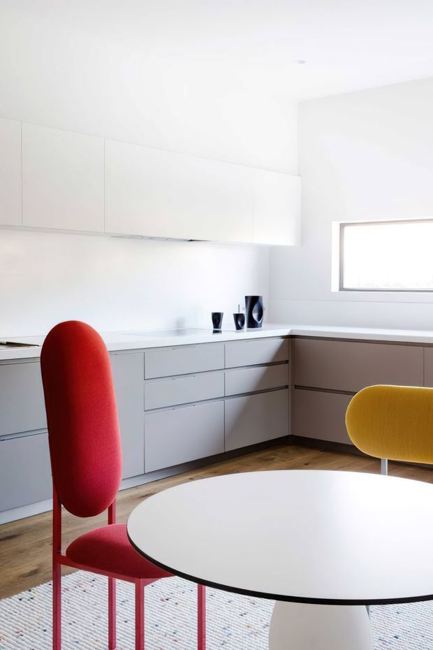 Bright minimalist kitchen and dining area with extensive matte white built-in cabinetry, pale benchtops, timber flooring, and a large rounded white dining table. A vivid red upholstered dining chair and mustard-yellow chair add bold colour accents, while sculptural dark decorative objects sit on the counter. A compact horizontal window provides natural light.