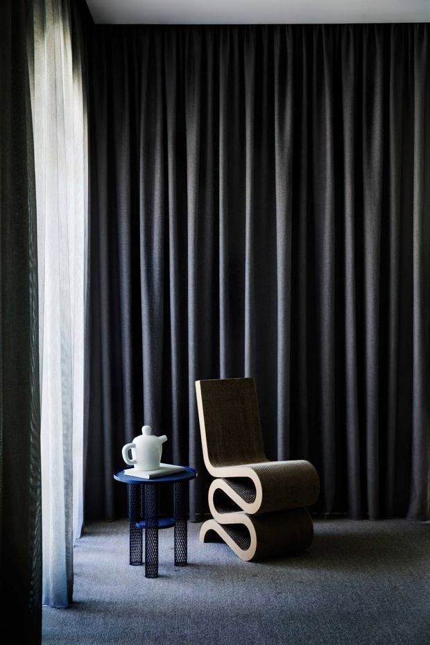 A moody interior vignette with full-height black drapery covering the back wall, a sheer curtain and narrow window at left, and dark carpeted flooring. A sculptural light-wood lounge chair sits beside a small round blue side table with a white ceramic teapot. The composition is minimalist and dramatic, with strong vertical folds and restrained furnishings.