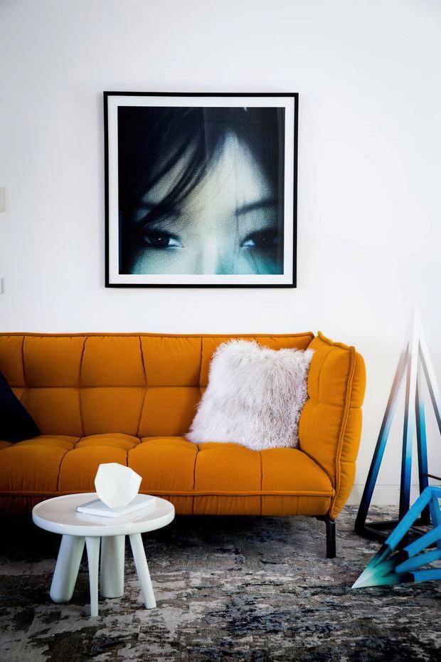 A styled living room vignette featuring a vivid mustard-orange tufted sofa, white shag cushion, round white side table, patterned grey rug, and bold contemporary artwork in a black frame. A sculptural floor lamp and turquoise geometric decor add graphic accents against white walls.