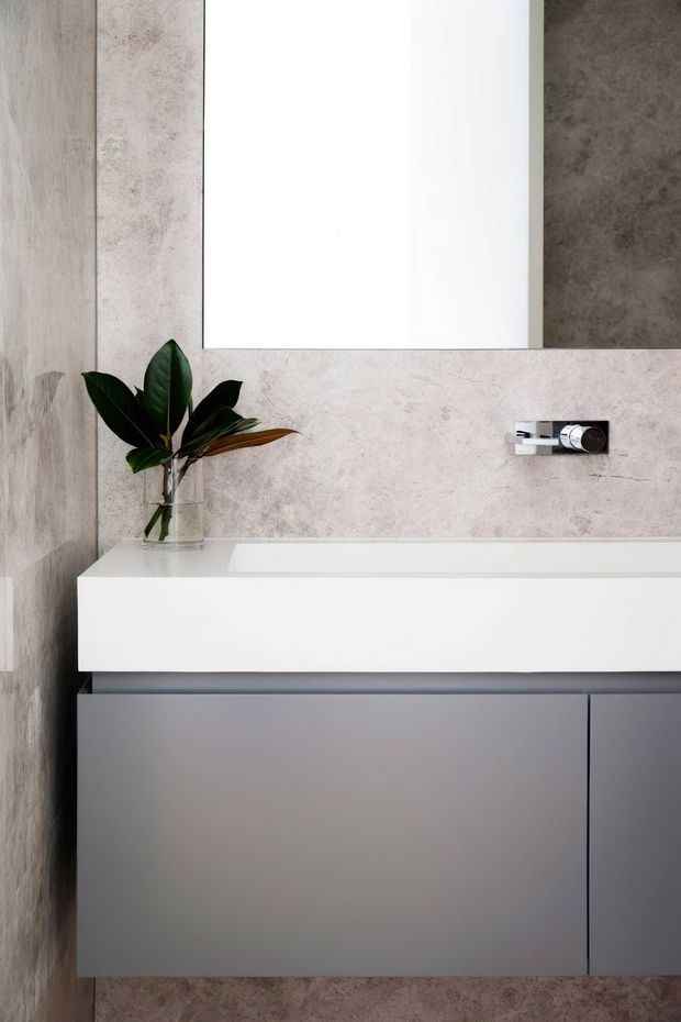 A close-up view of a contemporary bathroom vanity featuring a wall-mounted grey cabinet, white integrated basin and benchtop, large frameless mirror, and wall surfaces clad in pale mottled stone-look tile. A chrome wall-mounted tap and a small glass vase with leafy foliage add detail.