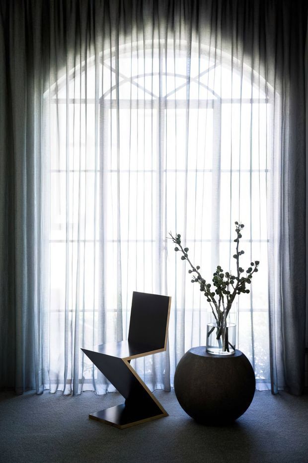 A moody, minimalist interior vignette featuring a sculptural black angular chair and a large rounded black pedestal or vessel with a tall branch arrangement. Floor-to-ceiling sheer curtains frame a large arched window, with soft diffused daylight creating a high-contrast, monochromatic atmosphere.