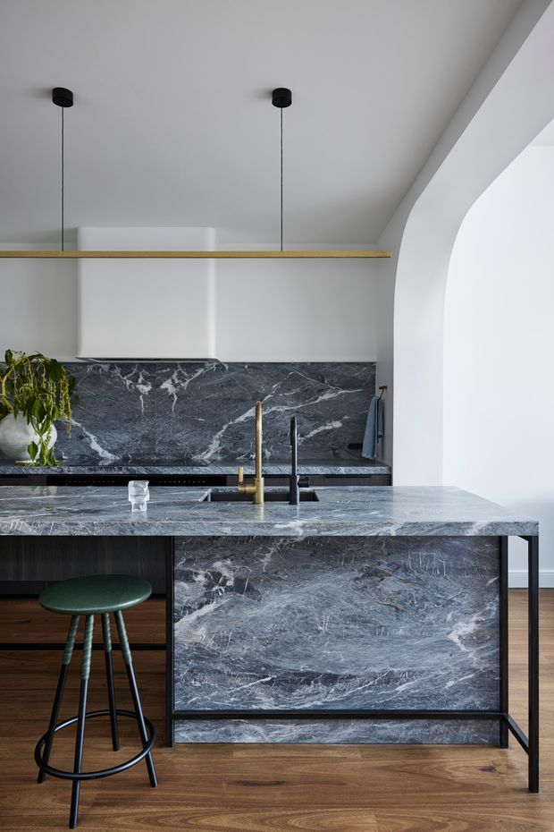 A refined contemporary kitchen centred on a large marble island with an integrated sink and brass and black taps. Dark veined marble continues across the splashback and island front, contrasted with dark cabinetry, warm timber flooring, white walls, and a slim timber open shelf. A green upholstered bar stool and pendant lights add restrained colour and function. A curved white architectural opening is visible on the right.