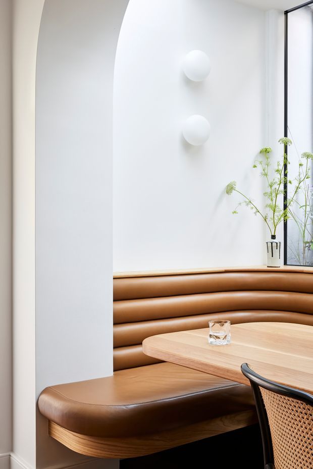 A refined dining nook with a curved built-in caramel leather banquette wrapping around a light timber table. The space has white plaster walls, a tall black-framed window, sculptural spherical wall lights, and delicate greenery, creating a clean contemporary interior with warm natural accents.