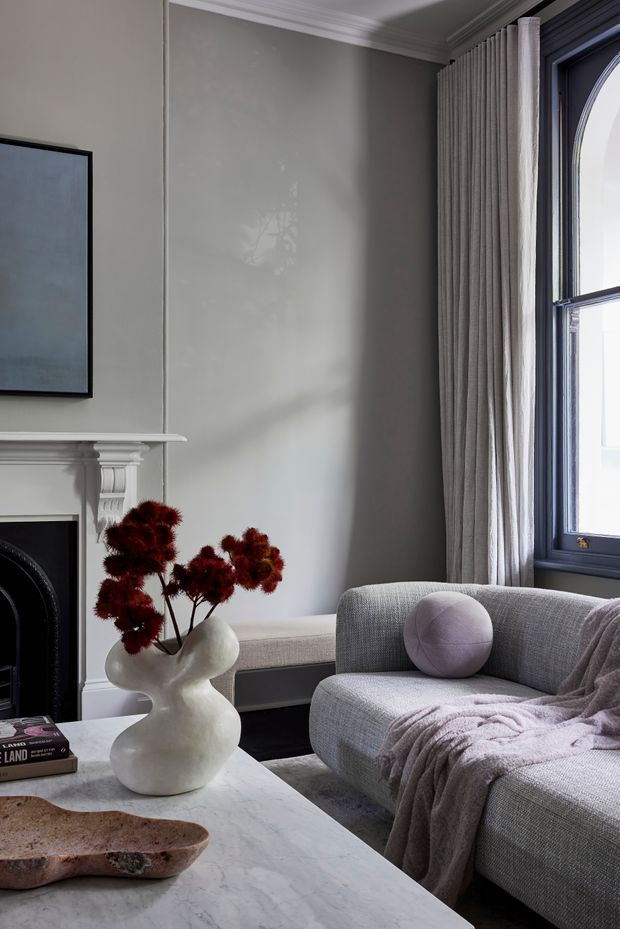 A refined living room with a grey upholstered curved sofa, soft mauve cushion and throw, a white marble coffee table, sculptural ceramic vase with dried red foliage, and a traditional fireplace with ornate mantel. Tall pleated curtains frame a dark-painted arched window, while neutral grey walls and classic cornice detailing create a calm, elegant atmosphere.