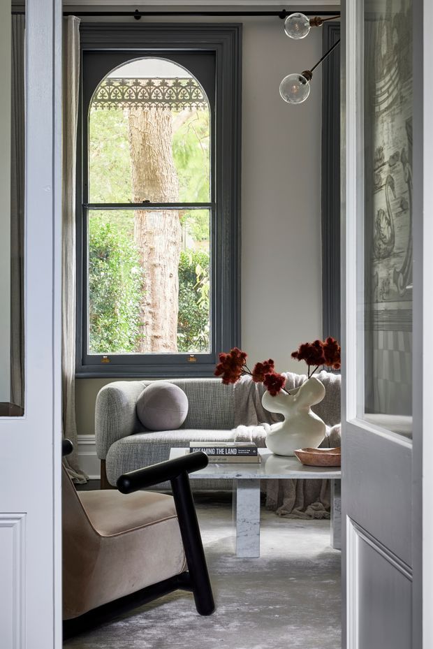 A refined sitting room viewed through partially open paneled doors. The room features a grey upholstered sofa with a round cushion, a sculptural white coffee table, a neutral armchair with a black angular frame, a soft grey rug, and dark grey arched window trim with decorative patterned glazing above the sash window. A sculptural wall light and floral arrangement add visual interest. The overall composition is elegant, layered, and softly lit.