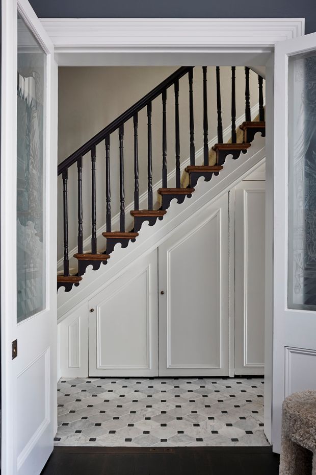 A refined entryway viewed through open glazed doors, featuring a staircase with dark turned timber balusters, dark handrail, warm timber stair treads, and decorative scalloped trim. White painted panelled joinery encloses the area beneath the stairs, while the floor has a patterned light stone or tile finish with small dark square insets. Ornate white ceiling mouldings and frosted decorative glazing reinforce the traditional character.