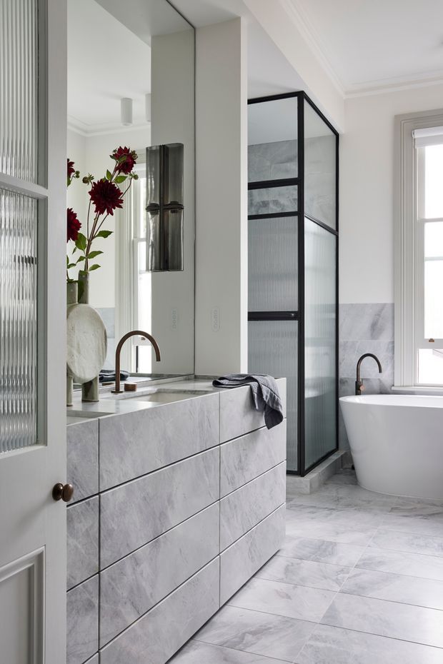 Bright, refined bathroom with a freestanding white bathtub, black-framed fluted-glass shower enclosure, marble-look tiled floor and walls, and a large floating vanity clad in pale grey veined stone. A mirrored wall, curved dark metal tapware, classic moulded cornices, and a window with a roller blind complete the space.