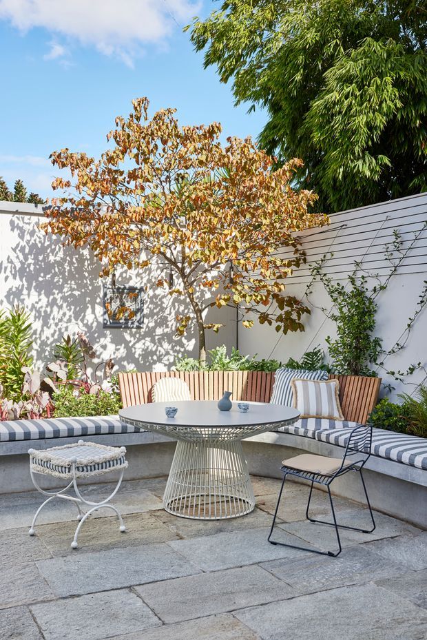 A bright, leafy courtyard patio with a curved built-in banquette upholstered in blue-and-white striped fabric, paired with a round outdoor table, wire-base chairs, and a small stool. Pale rendered walls, horizontal timber screening, climbing plants, mature trees, and large grey stone paving create a relaxed garden setting.