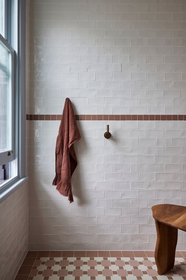 A bright, narrow tiled interior featuring a full-height white rectangular tile wall with a horizontal terracotta-brown accent stripe. A rust-coloured towel hangs from a small dark metal wall hook, while patterned floor tiles combine white, terracotta, and muted mauve squares. A timber stool or small side table sits in the lower right, and a tall window provides natural light on the left.