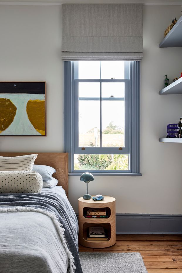 A bright, carefully styled bedroom corner featuring a timber bed with layered neutral bedding, a small rounded timber bedside table with open shelving, and a sculptural table lamp. A large sash window with blue-grey timber trim and a Roman blind provides natural light. Built-in floating shelves display books and small objects, while a framed abstract artwork hangs on the pale wall. Timber floorboards and a grey rug complete the calm, contemporary setting.