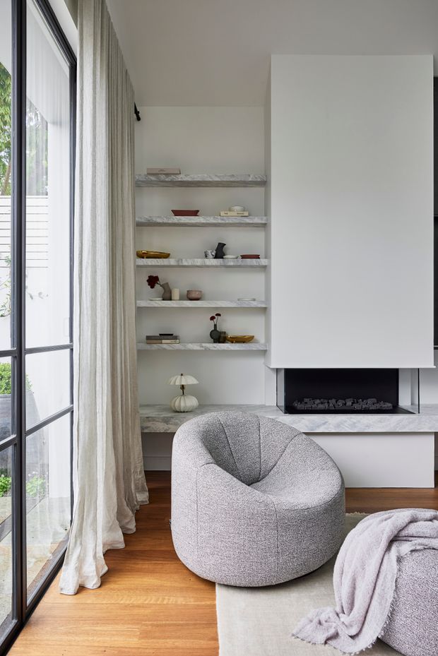 A refined living room with a sculptural boucle-style lounge chair, a matching partially visible sofa, pale rug, timber flooring, full-height sheer curtains and black-framed glazing. A minimalist white fireplace wall with a broad marble-look hearth and recessed open display shelving forms the main architectural feature. The palette is soft white, grey, pale timber and muted blush, with carefully styled objects on the shelves.