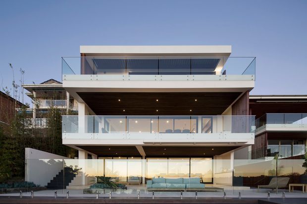 Contemporary multi-level residential facade viewed from the rear, featuring extensive floor-to-ceiling glazing, broad projecting balconies with clear glass balustrades, white rendered horizontal volumes, and warm timber-lined soffits. The lower level opens onto a pool terrace with outdoor seating and views beyond. The building has a flat roof and strong cantilevered forms.
