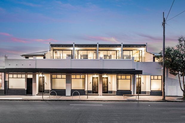 Street-facing view of a contemporary multi-storey building at dusk, with a symmetrical facade, white rendered walls, dark horizontal bands, extensive upper-level glazing, balconies or terraces, and ground-floor commercial-style entries. Utility poles and a paved street occupy the foreground.