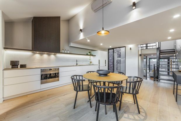 Open-plan contemporary kitchen and dining area with white handleless cabinetry, a dark wood feature panel, linear tiled splashback, light timber flooring, and a round timber dining table with black spindle-back chairs. Recessed downlights and a central pendant illuminate the space, while a glazed and metal-framed stairway is visible to the right.