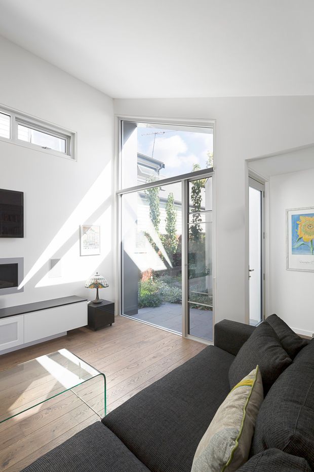 Bright contemporary living room with white walls, pale timber flooring, a large full-height glazed door opening to a small courtyard, and a narrow high window. A dark charcoal sectional sofa, minimalist white media cabinetry, glass coffee table, and small lamp furnish the room. Strong sunlight creates diagonal shadows across the floor and walls.