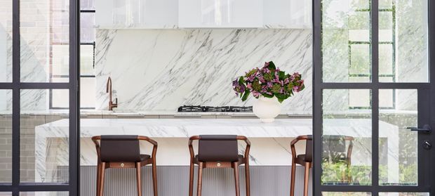 A bright contemporary kitchen viewed through black-framed glazed doors. The space features a large white marble-look island with integrated seating, a matching veined stone splashback, white upper cabinetry, a dark cooktop, and warm timber-and-leather bar stools. Greenery is visible beyond the glazing, adding a natural element.