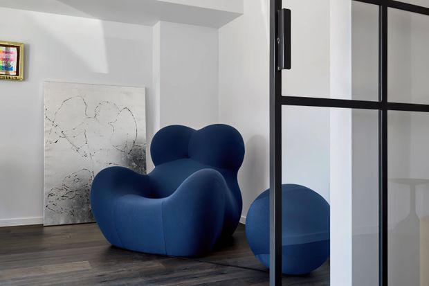 A contemporary interior vignette featuring a sculptural deep-blue upholstered lounge chair and matching rounded ottoman on dark timber flooring. White walls, abstract artwork, and a black-framed glazed partition create a minimalist, gallery-like setting.