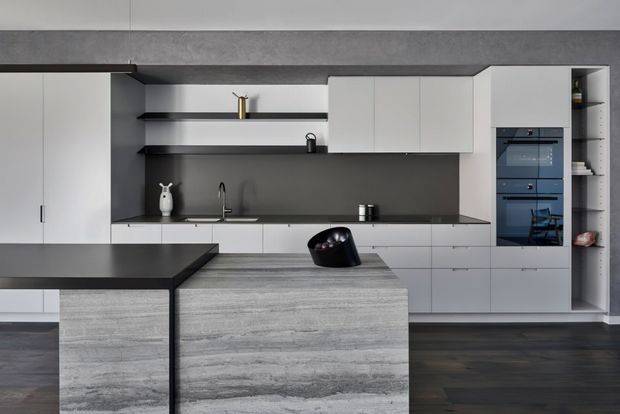 Contemporary minimalist kitchen with matte white handleless cabinetry, dark grey splashback and benchtops, open black shelving, integrated appliances, and a large grey stone island. Dark timber flooring and restrained styling create a refined monochromatic interior.