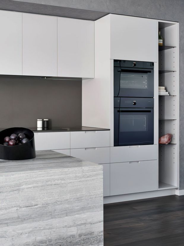 Contemporary minimalist kitchen featuring matte white handleless cabinetry, integrated double wall ovens, dark grey splashback and benchtop, open pantry shelving, a large pale stone island, and dark timber flooring. The kitchen has a clean, restrained monochromatic palette with strong horizontal and vertical lines.
