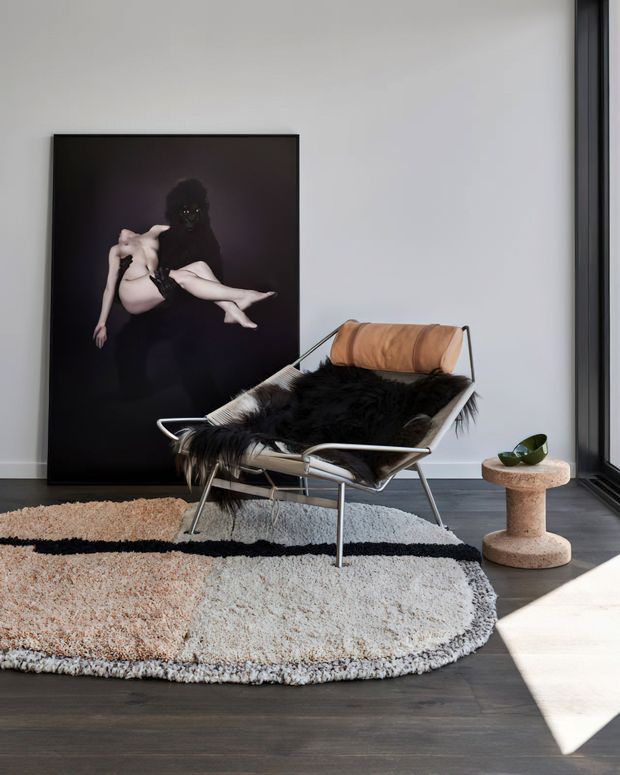 A refined modern interior vignette featuring a black leather-and-metal sling lounge chair layered with a dark shag throw, positioned beside a small rounded stone side table with a potted plant. A large monochrome artwork leans against a pale wall, while a textured neutral area rug sits over dark timber flooring. Strong sunlight enters through tall black-framed glazing, creating a calm, high-contrast setting.