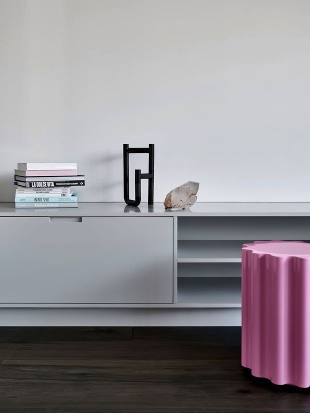 Minimalist interior vignette featuring a sleek grey built-in sideboard with closed cabinetry and open shelving, styled with books, a black sculptural object and a stone ornament. A bright pink fluted stool stands in front on dark timber flooring against a plain light wall.