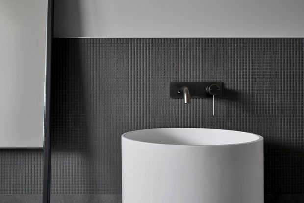 Close-up of a contemporary bathroom vanity featuring a white cylindrical vessel basin against a dark micro-mosaic tiled wall. A wall-mounted dark metal tap and control are centered above the basin, with a dark-framed edge and partial pale wall visible at left.