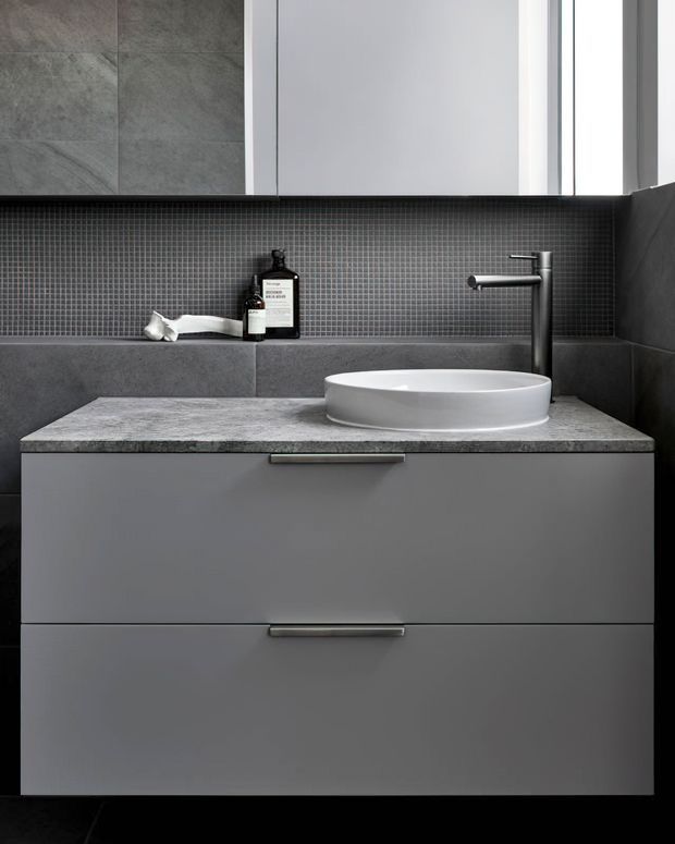 Contemporary bathroom vanity with a wall-mounted grey drawer cabinet, mottled grey stone benchtop, white round vessel basin, dark grey micro-mosaic tiled splashback, charcoal wall tiles, and a minimalist black tap. A large mirror and narrow window add brightness to the otherwise monochromatic space.