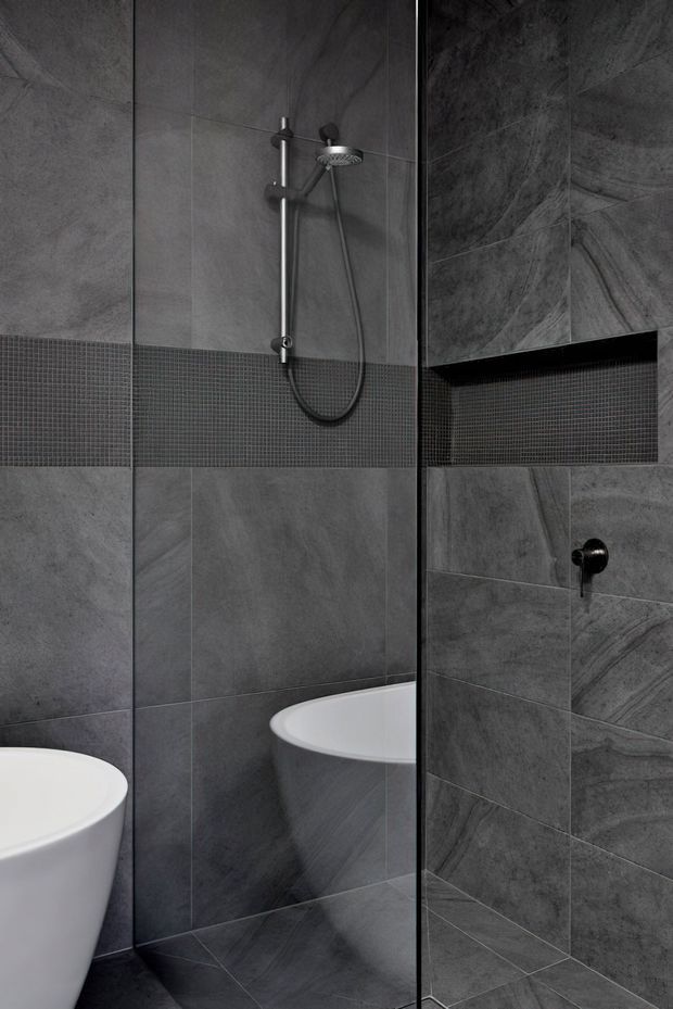 A compact contemporary bathroom with a frameless glass shower enclosure, dark grey stone-look wall and floor tiles, a horizontal band of small mosaic tiles, matte black shower fittings, a recessed wall niche, and a partially visible white freestanding bathtub.