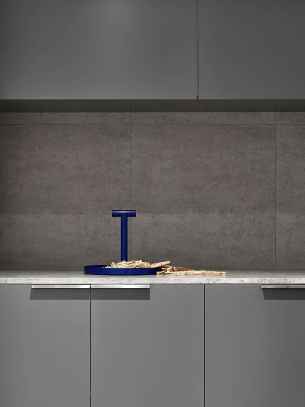 Close-up of a minimalist kitchen or utility joinery wall with dark grey flat-panel cabinetry, a large grey textured slab splashback, and a pale stone-look benchtop. A distinctive deep-blue tap and shallow blue vessel sit on the counter, with small timber pieces nearby. The composition is clean, linear, and highly restrained.