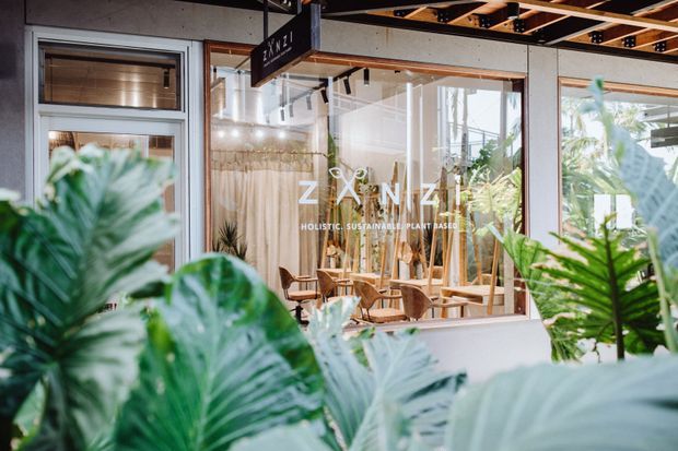 Exterior view through lush tropical foliage toward the glazed storefront of ZENZI, a holistic sustainable plant-based venue. The facade features large timber-framed windows, a light rendered wall, and a timber-and-metal pergola or canopy overhead. Indoor café-style seating and abundant plants are visible through the glazing.