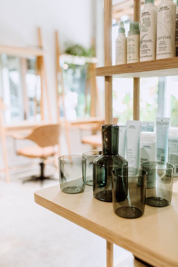 Bright, airy salon or retail display interior featuring pale timber shelving and a matching counter, with Aveda hair-care products, glass bottles, and tumblers arranged in the foreground. Large mirrors and windows create soft reflections and a light, natural atmosphere; salon seating is visible in the background.