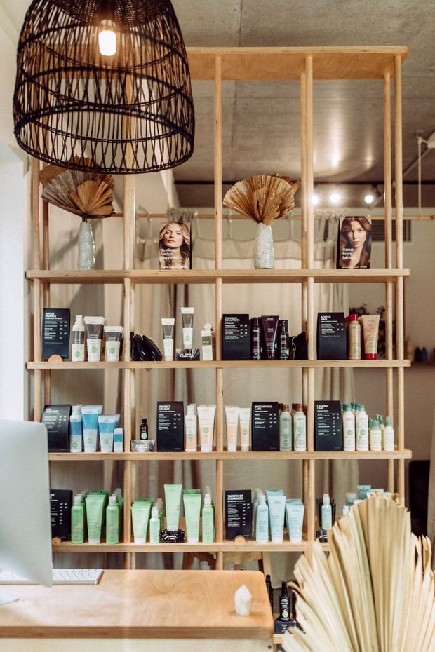 A warmly lit retail or beauty salon display area with an open timber shelving unit stocked with skincare products, brochures, and decorative dried-leaf arrangements. A large woven pendant light hangs above, with mirrors and a concrete-look ceiling contributing to the natural, tactile interior.