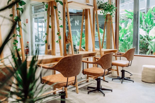 Bright plant-filled commercial interior with a row of timber-framed workstations or styling stations, integrated mirrors, and warm brown upholstered swivel chairs. Large windows overlook dense tropical greenery, creating an airy, biophilic workspace or salon-like environment. The floor is light-toned and the fit-out uses extensive natural timber and indoor planting.