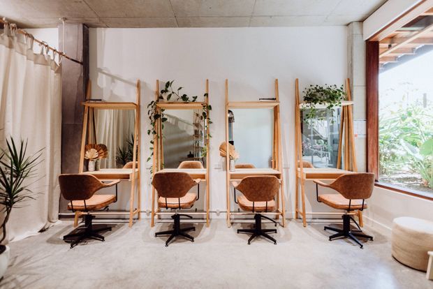 A bright, minimalist salon or beauty workspace with four individual styling stations arranged along a white wall. Each station has a timber-framed mirror, narrow timber shelf, potted greenery, and a brown upholstered swivel chair. The room features a polished concrete ceiling and floor, a large timber-framed window, sheer curtains, and abundant natural light.