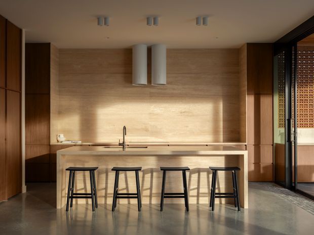 A warm, minimalist kitchen with a long pale timber island and integrated sink, four black stools, full-height timber cabinetry, matching timber wall panelling, cylindrical rangehoods, and polished concrete flooring. Strong sunlight enters through dark-framed glazing and a patterned brick screen at the right.